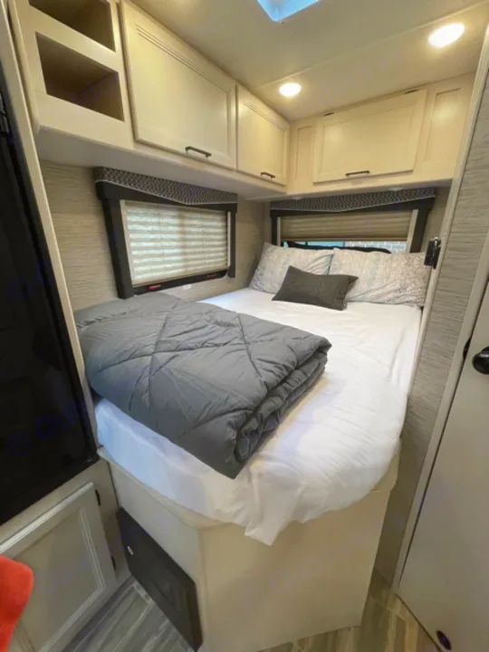 Clean and modern RV rental bedroom featuring a comfortable bed, overhead cabinets, and windows with blinds.. Thor Motor Coach Four Winds 2022