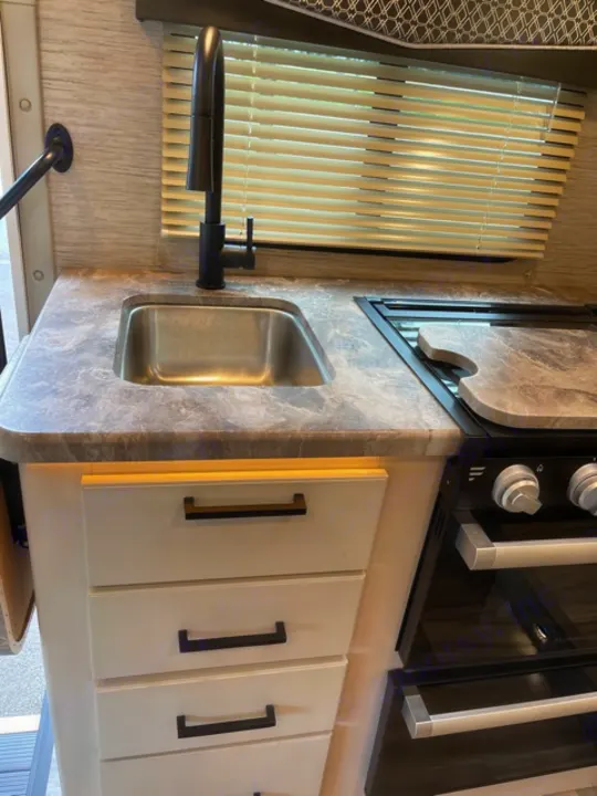 Modern RV rental kitchen featuring a stainless steel sink, sleek black faucet, window with blinds, and white cabinets.. Thor Motor Coach Four Winds 2022