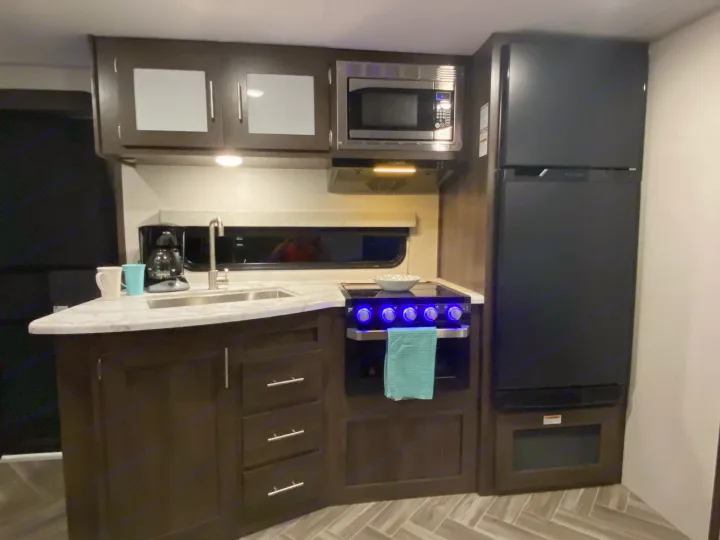 Compact RV rental kitchen features dark wood cabinets, a sink, microwave, oven with blue knobs, and a large black refrigerator.. K-Z Manufacturing Sportsmen 2021