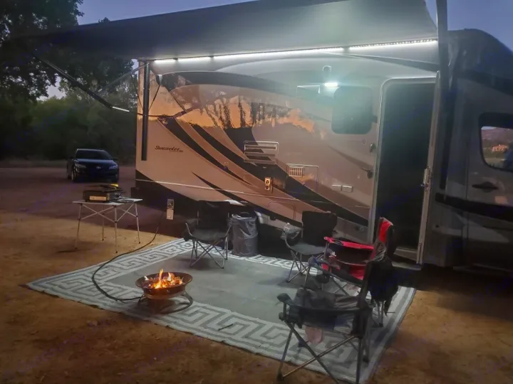 An evening view of a well-lit RV rental campsite, featuring an awning, fire pit, and comfortable outdoor seating.. Forest River Sunseeker 2017