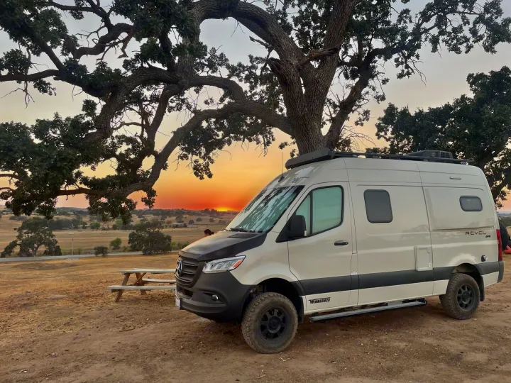 Experience an RV rental: a beige Winnebago Revel 4x4 camper van parked under a sprawling tree at sunset.. Winnebago Revel 2021