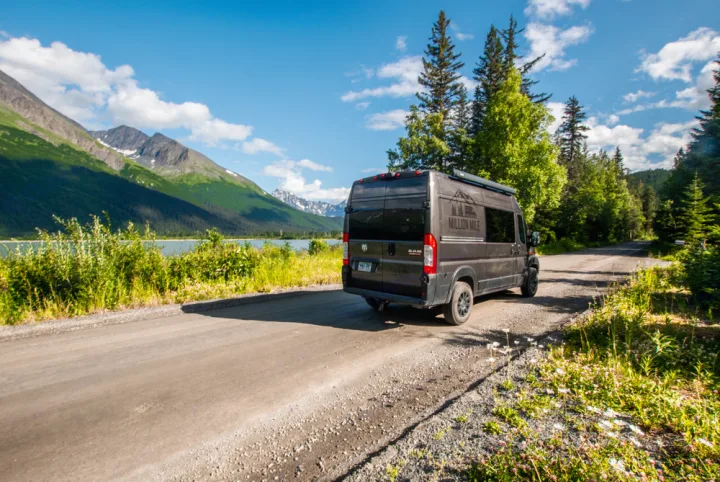 Experience nature with an RV rental! A dark camper van is parked on a dirt road by a serene lake & majestic snow-capped mountains.. Ram Promaster 2020