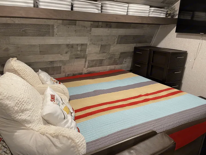 Cozy RV rental bedroom featuring a colorful striped bed, rustic wood wall, overhead storage, and dresser.. Coleman Other 2022