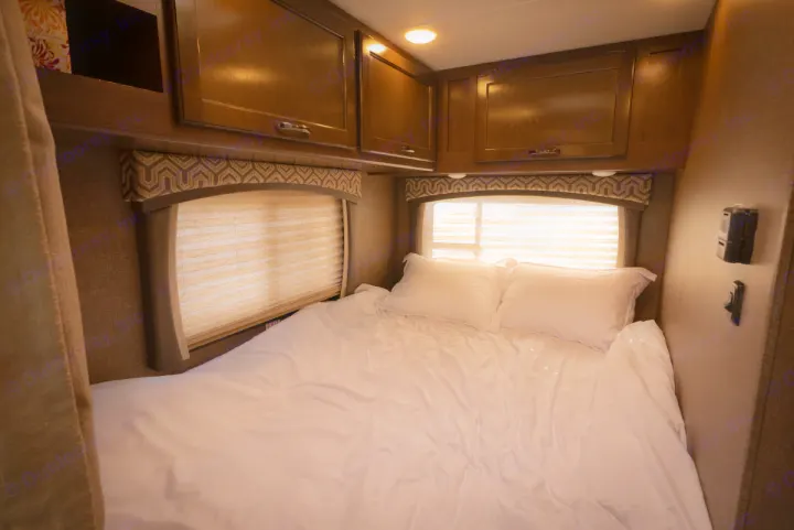 Experience comfort in this RV rental bedroom: a neat bed with white sheets, two pillows, windows with blinds, and wood cabinets.. Thor Motor Coach Chateau 2019