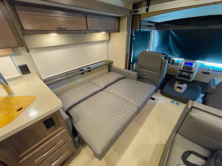 Modern RV rental interior featuring a grey convertible sofa bed, compact kitchen, and cockpit view, ideal for travel.. Winnebago Adventurer 2022
