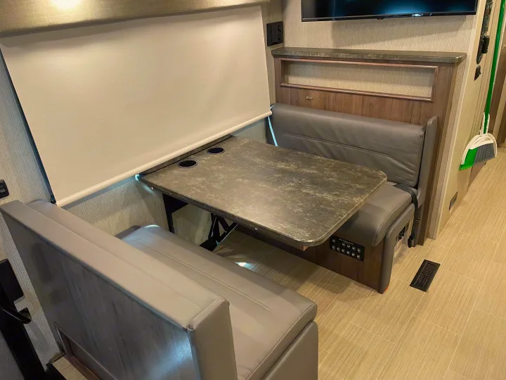 Cozy RV rental dinette featuring comfy gray benches, a table with cupholders, and a roll-down window blind.. Winnebago Adventurer 2022