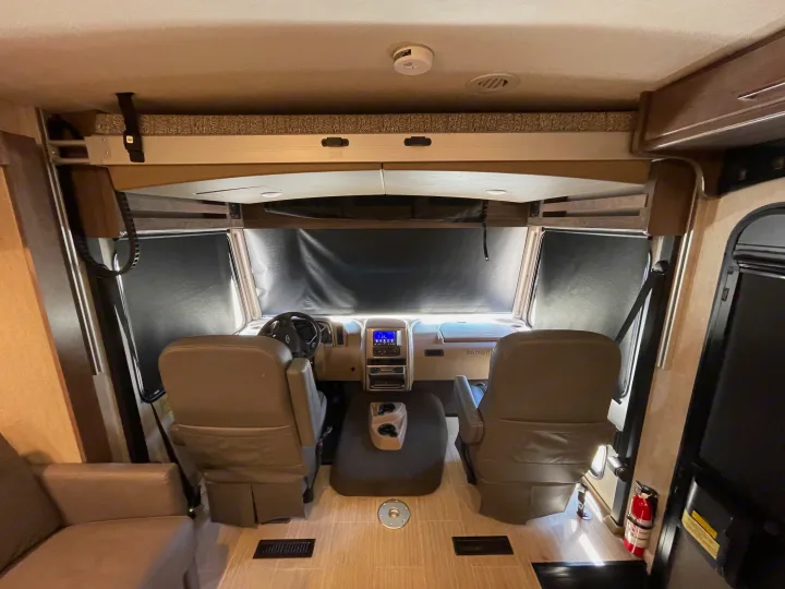 Front view of an RV rental's driving area, featuring blackout window covers, two seats, dashboard, and overhead bed.. Winnebago Adventurer 2022