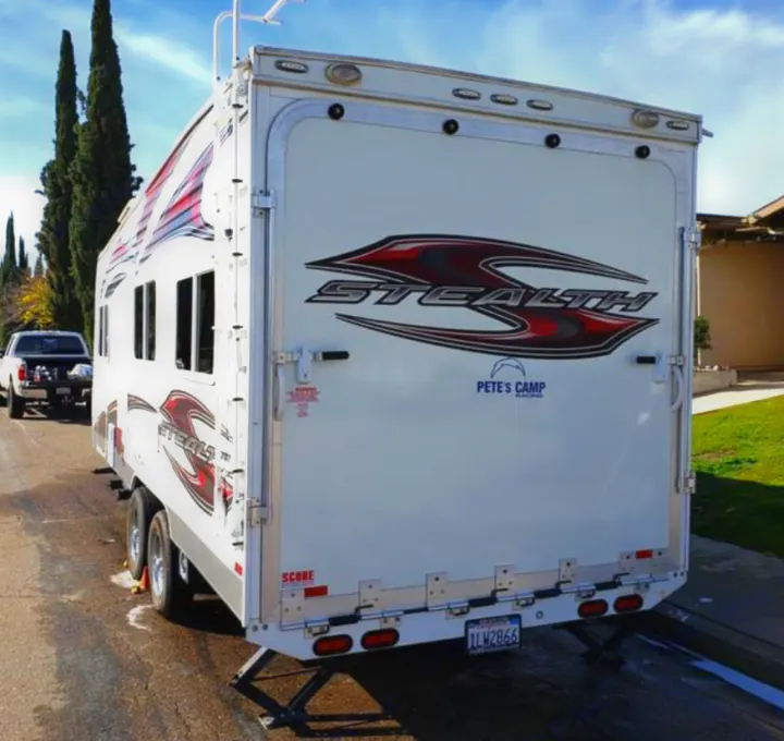 Rear view of a white "Stealth" toy hauler RV trailer, featuring its ramp door and stabilizing jacks, parked on a street. Great for an RV rental.. Forest River Stealth 2012