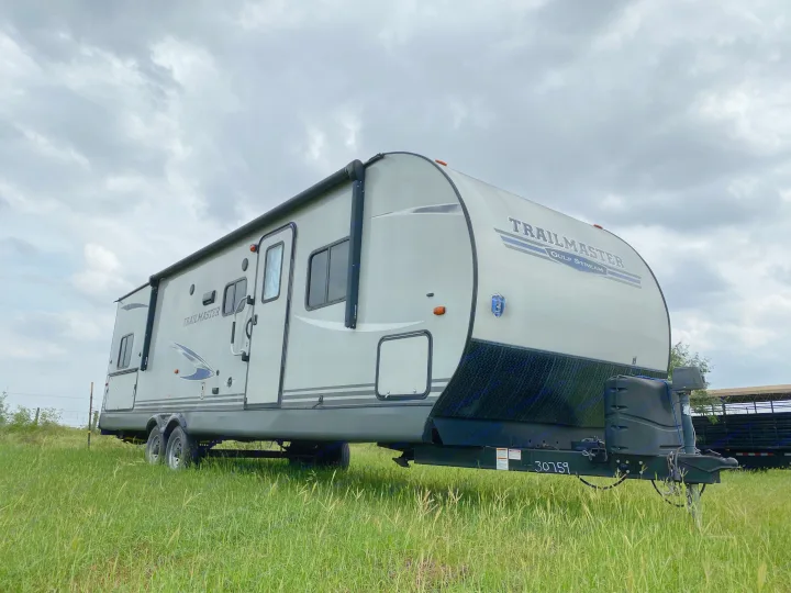 A gray Trailmaster Gulf Stream travel trailer, perfect for an RV rental, stands in a green field under a cloudy sky.. Gulf Stream Trailmaster 2020