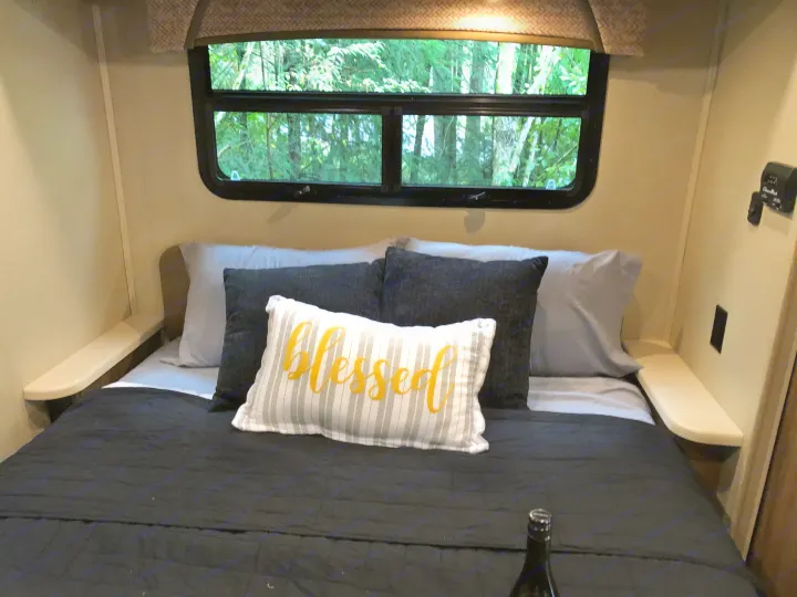 Cozy RV rental bedroom with a bed, "Blessed" pillow, and a window overlooking lush green trees, plus side shelves.. Jayco Greyhawk 2018
