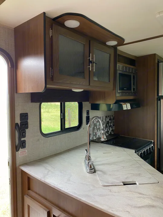 Modern RV rental kitchen with wood cabinets, light countertops, sink, microwave, and stove.. Gulf Stream Trailmaster 2020