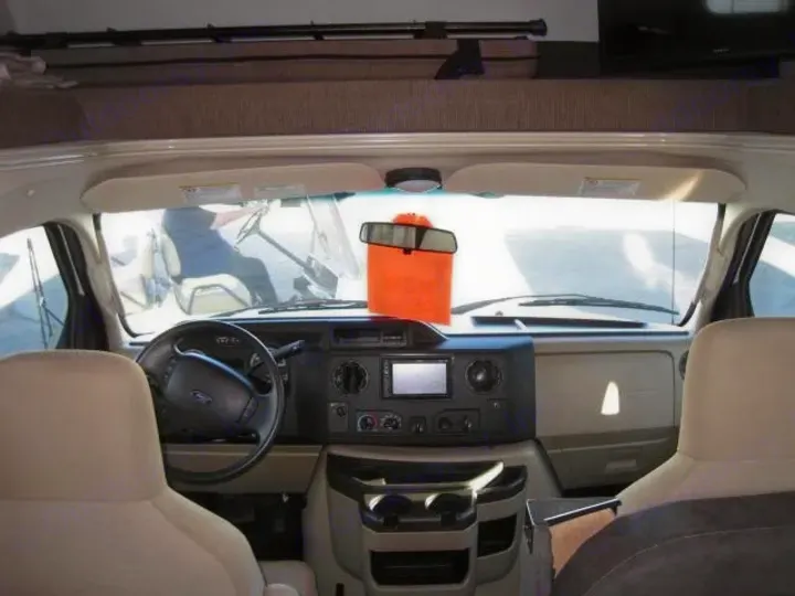Inside an RV rental: the spacious cockpit with dashboard, steering wheel, and wide front windshield, viewed from the living area.. Coachmen Leprechaun 2021
