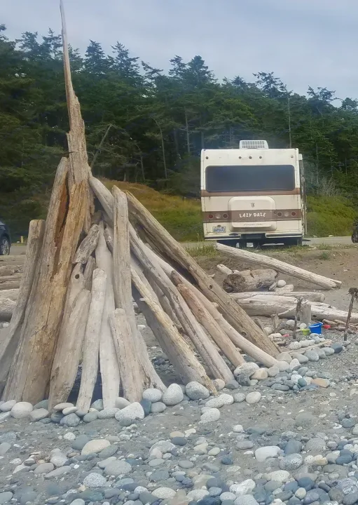 A large driftwood teepee on a rocky beach with a "LAZY DAZE" rv rental parked on the road, backed by green trees.. Lazy Daze 22 1987