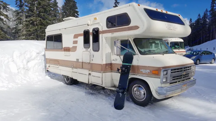 White and brown Lazy Daze Chevy RV rental in a snowy landscape with a snowboard. Perfect for winter travel.. Lazy Daze 22 1987