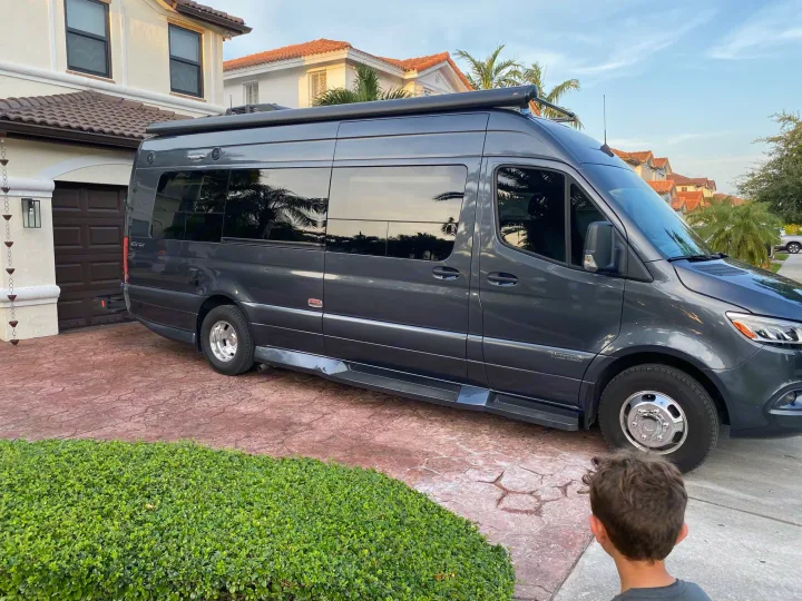 A dark gray **RV rental** van is parked in a residential driveway. A child looks on, seeing the sleek Sprinter.. Winnebago Era 2021