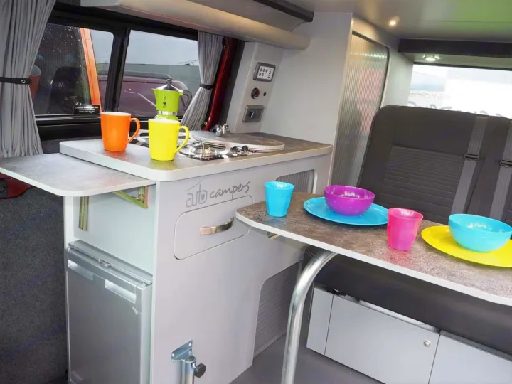 RV rental's compact kitchen and dining area: stove, sink, fridge, and colorful dinnerware, ideal for travelers.. Ford Transit Custom 2+2 berth Pop Top, Manual 2022