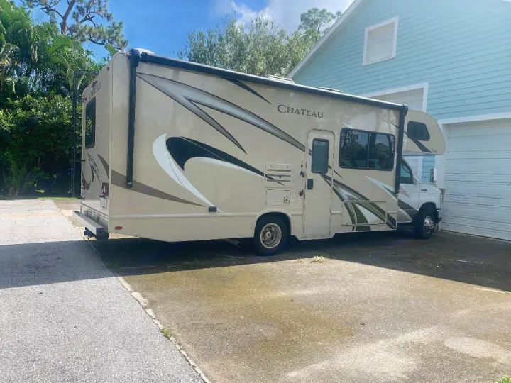 A beige Chateau RV rental is parked on a sunny driveway next to a light blue house, ready for adventure.. Thor Motor Coach Chateau 2020