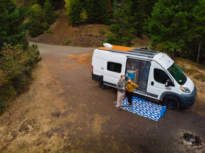 Two travelers pose with their white camper RV rental van on a dirt road in a wooded area.. Dodge RAM Promaster 2500 159' Wheelbase 2017