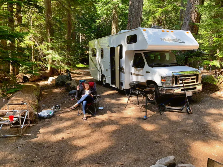 A woman relaxes by her rv rental in a peaceful forest campsite, complete with a dog, grill, and electric scooter.. Thor Majestic 2015