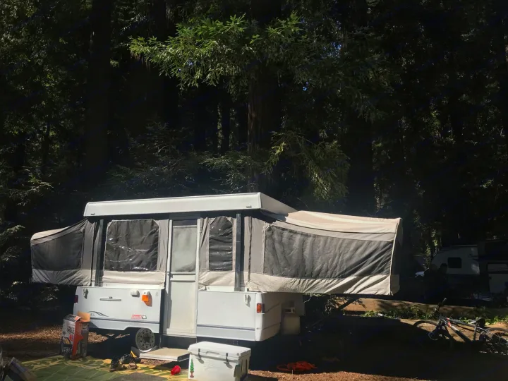 An RV rental pop-up camper fully set up at a sunny, shaded forest campsite with outdoor gear & bikes.. Coleman Grand Tour Carmel 2003