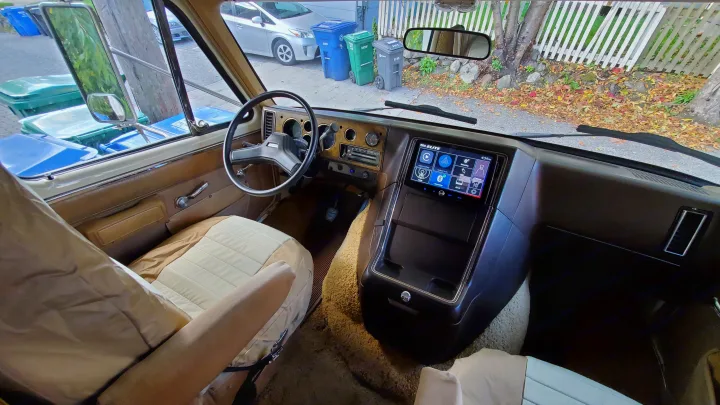 Interior of an RV rental: vintage dashboard, modern display, tan seats, and the view out the windshield with yard waste bins.. Lazy Daze 22 1987