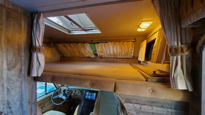 Inside an RV rental: a spacious cab-over bunk with skylight, windows, curtains, and the driver's area below.. Lazy Daze 22 1987