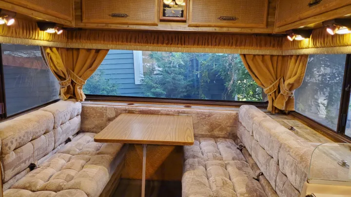 Interior of an RV rental: a cozy dinette with plush benches, a wooden table, and large windows revealing lush greenery outside.. Lazy Daze 22 1987