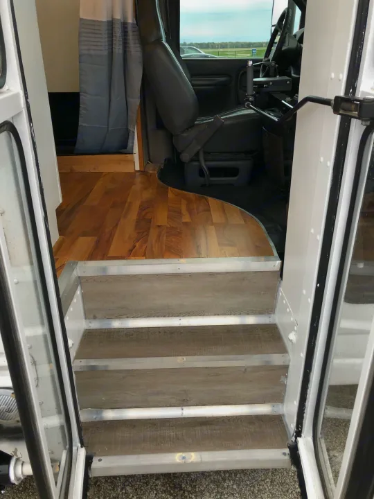 RV rental interior: Steps with wood-look treads lead to laminate flooring and the front driver's area of this converted van.. Chevy Express 3500 Mini Bus 2002