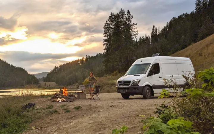 A woman enjoys lakeside camping in a white Mercedes Sprinter RV rental. A campfire glows at sunset, surrounded by mountains.. Mercedes-Benz Mercedes Sprinter 2500 2015