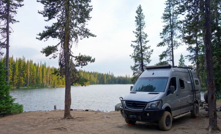 Lakeside camping with an RV rental. A gray custom camper van is parked on dirt next to a calm lake and dense forest.. Winnebago Revel 2019