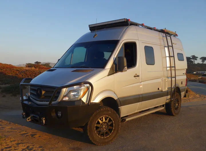 Custom silver Mercedes Sprinter RV rental parked outdoors. This rugged adventure camper van features off-road tires, a winch, and roof rack.. Winnebago Revel 2019