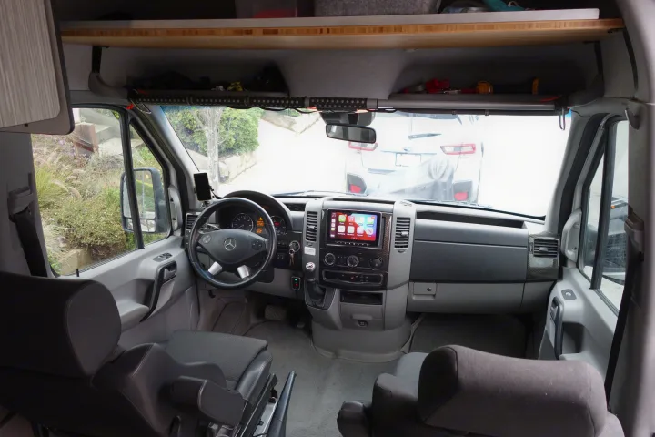 Front cabin of an RV rental showing the steering wheel, dashboard, infotainment display, and comfortable front seats.. Winnebago Revel 2019