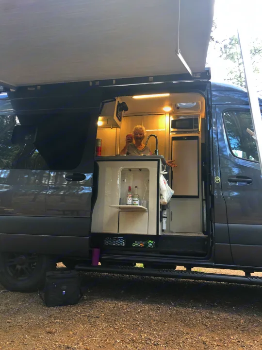 Happy woman in her spacious RV rental's mobile kitchen, complete with sink, microwave, fridge, and awning for camping enjoyment.. Mercedes-Benz 4x4 Sprinter mwb 2021