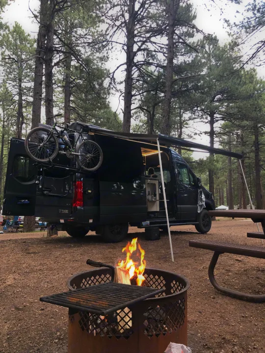 Perfect **RV rental** setup: black camper van with bike rack, awning extended. Campfire burns beside a picnic table in a pine forest.. Mercedes-Benz 4x4 Sprinter mwb 2021