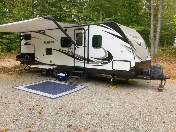 RV rental: A modern white travel trailer with an extended awning is parked on a gravel campsite with an outdoor rug.. Keystone Passport 2017