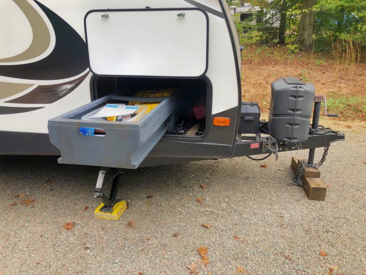 Front of an RV rental at a campsite. Its exterior storage drawer is pulled out, showing gear; leveling jacks deployed.. Keystone Passport 2017
