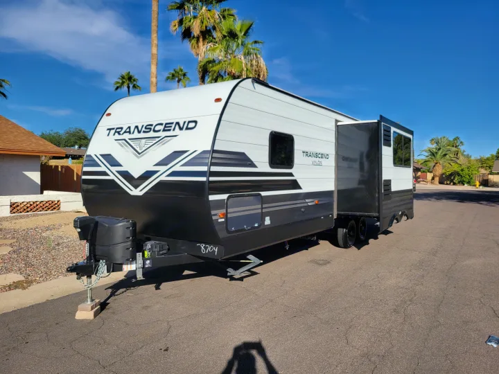 A white and black Transcend XPLOR travel trailer RV rental, with a slide-out, parked on asphalt with palm trees.. Grand Design 265bh 2021