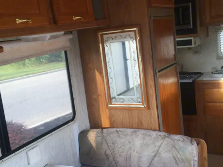 Inside a comfortable RV rental: wood cabinets, mirror, patterned sofa, and kitchen appliances. Window shows a road view.. Gulf Stream Conquest 1998