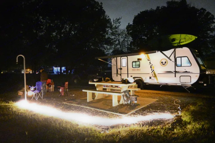 Bright lights illuminate this RV rental campsite at night, featuring a travel trailer, picnic table, and person camping.. Forest River R-Pod 2020