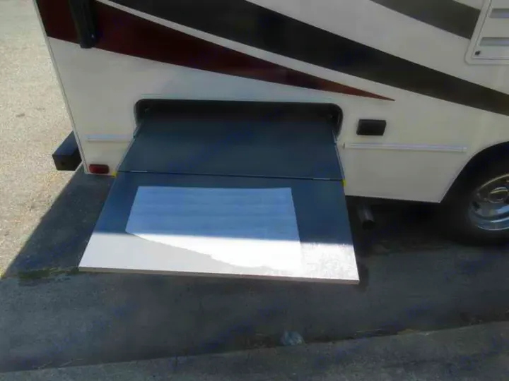 An RV rental's side compartment reveals a grey, two-section pull-out storage tray extended onto asphalt.. Coachmen Freelander 2019