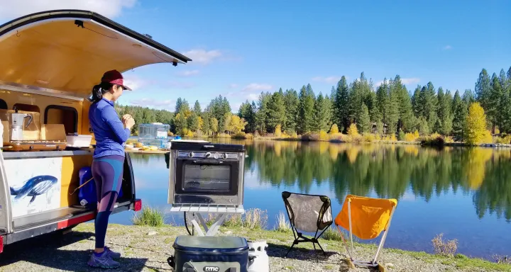 Lakeside cooking setup: A woman preps food at a teardrop RV rental, with a scenic lake & autumn trees.. Other Other 2011
