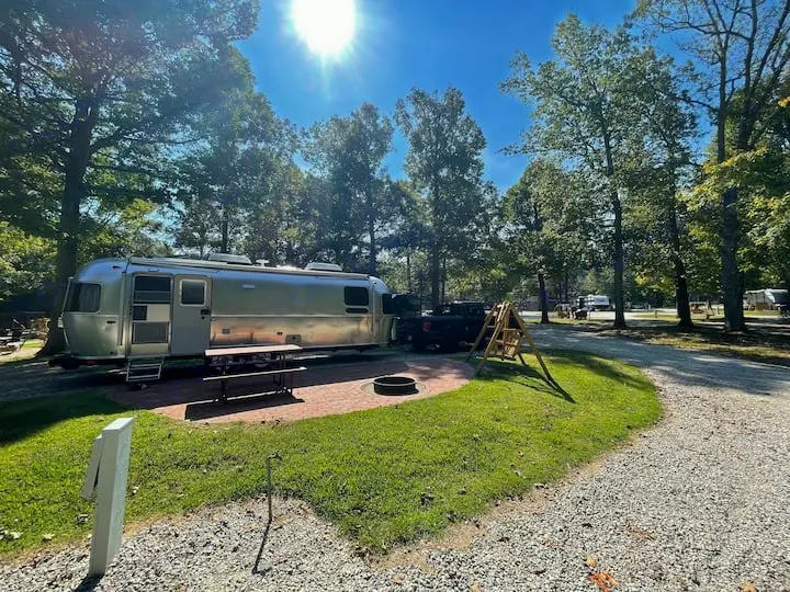 Serene campground with an Airstream RV rental featuring a patio, picnic table, and fire pit under sunny trees.. Airstream Globetrotter 2022