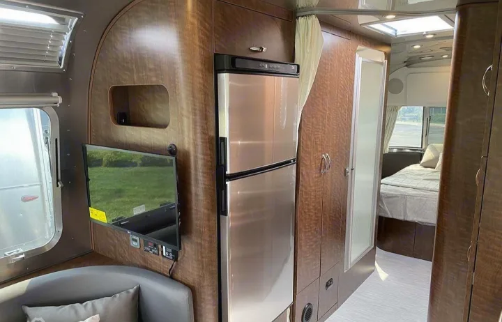 A detailed view inside a modern RV rental, showing its stainless steel fridge, TV, wood cabinetry, and bedroom.. Airstream Globetrotter 2022
