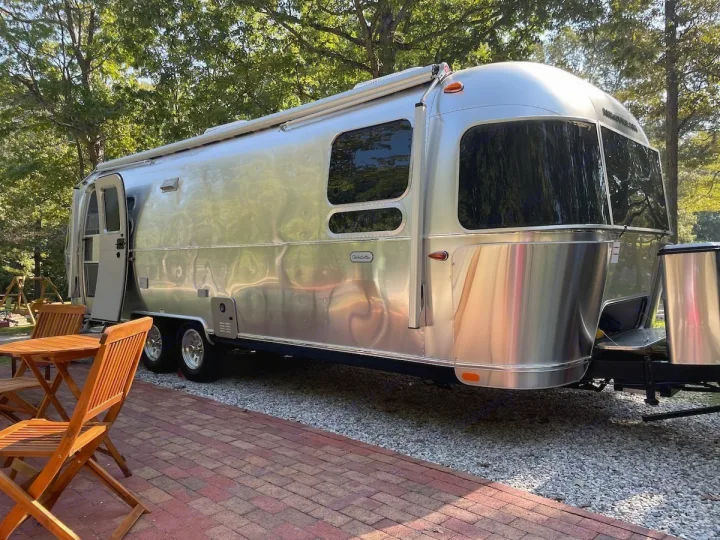 This Airstream travel trailer, ideal for an RV rental, is parked beside an outdoor patio with wooden furniture.. Airstream Globetrotter 2022