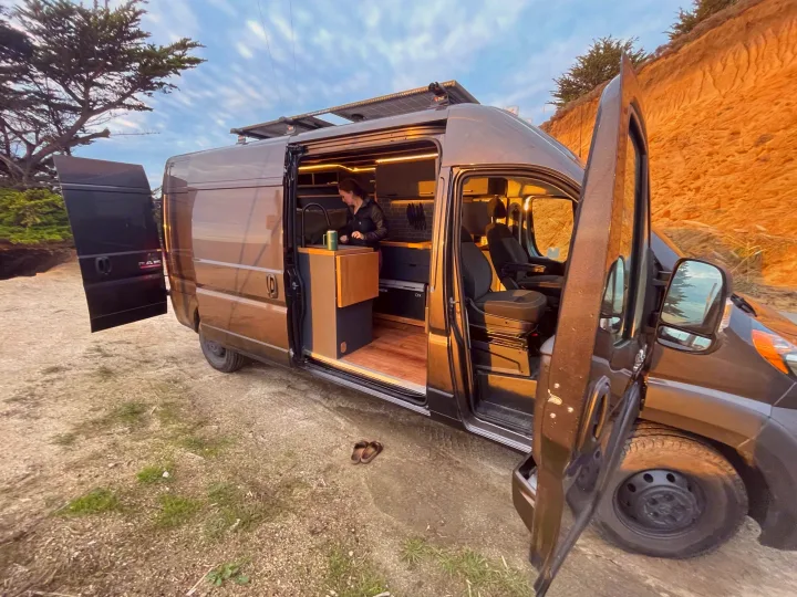 Explore in comfort with this RV rental! Woman makes coffee in a modern camper van kitchen, doors open to nature.. Ram Promaster 2021