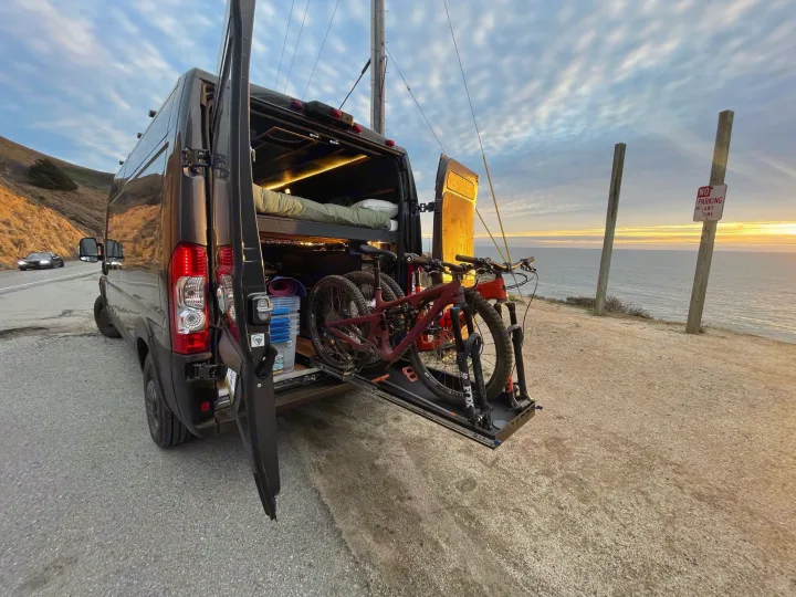 An RV rental, a converted van, is parked along a scenic ocean cliffside road at sunset, showcasing interior bed & bikes.. Ram Promaster 2021