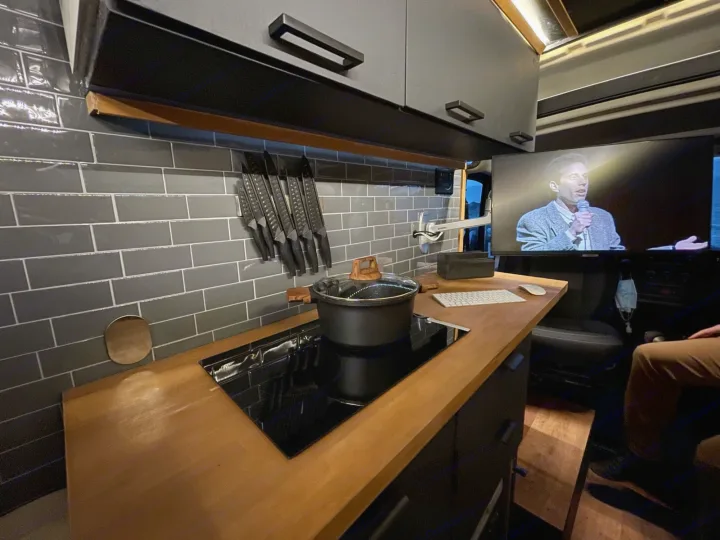 Modern RV rental kitchen with wood counters, an induction cooktop, and a TV showing a speaker. Practical and cozy.. Ram Promaster 2021