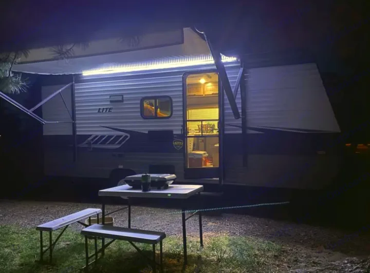 Cozy night camping setup with an RV rental. Lit awning and interior lights illuminate the outdoor table, grill, and benches.. Crossroads Zinger 2018