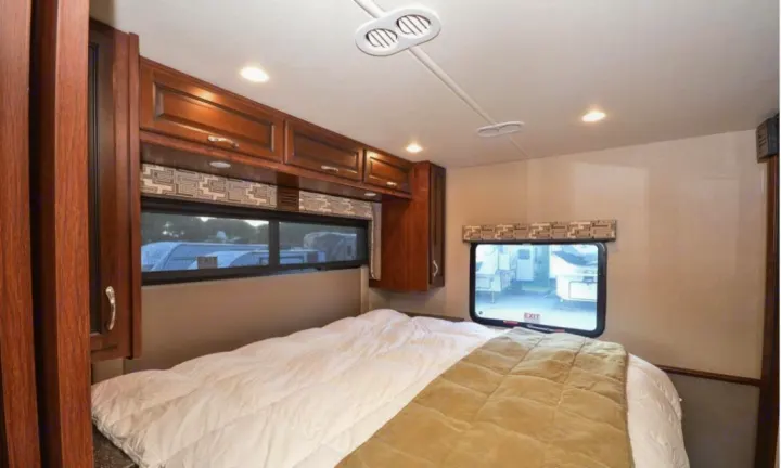 Interior view of an RV rental bedroom showing a bed with white and tan covers, wooden cabinets, and windows.. Fleetwood Other 2017