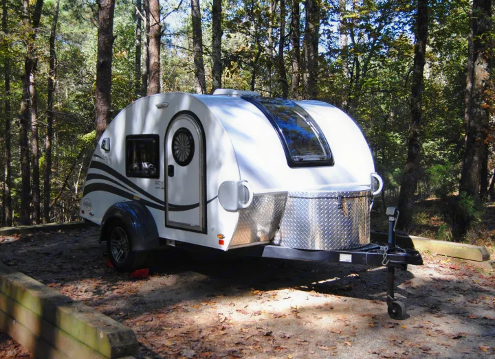 A white teardrop RV rental parked on a leaf-covered campsite, nestled among tall trees in a sunny forest.. nuCamp T@G 2021